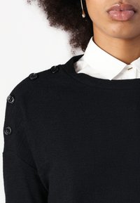 Black knitted sweater with a round neckline and button accents on the shoulder. Layered over a white collared shirt. Smooth texture.