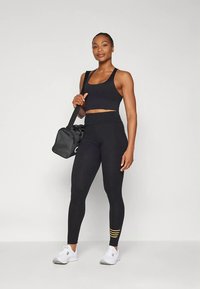 Μαύρο αθλητικό crop top και κολάν με ψηλή μέση και κίτρινες ρίγες. Λευκά αθλητικά παπούτσια και μαύρη αθλητική τσάντα με ιμάντα ώμου.