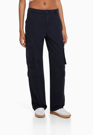 Osoba ubrana w granatowe spodnie cargo z bocznymi kieszeniami oraz białe sneakersy z brązowymi podeszwami, stojąca na białym tle.