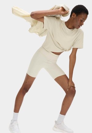 Person i beige kort t-shirt og shorts strækker med den ene hånd på knæet, holder jakke over skulderen, iført hvide sneakers og sokker.