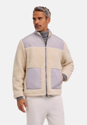 Mann trägt eine hellbeige Sherpa-Fleecejacke mit grauen Stoffeinsätzen und zwei Vordertaschen und schaut nach links.