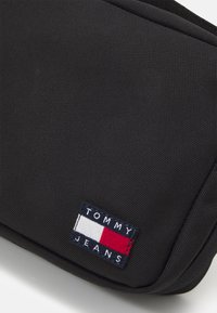 Svart tygväska med en strukturerad yta. Har en rektangulär Tommy Jeans-logotyp i rött, vitt och blått. Med dragkedjestängning.