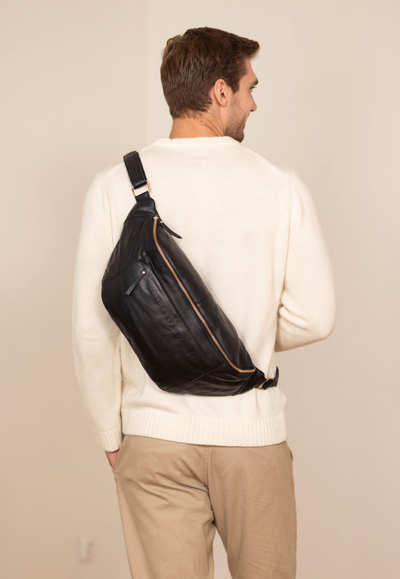Bolso de cuero negro en forma curva, con detalles en dorado en la cremallera y una correa de ajuste única, llevado en la espalda sobre un fondo claro.
