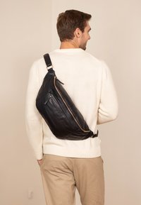 Schwarze Lederslingtasche mit einer geschwungenen Form, goldenen Reißverschlussakzenten und einem einzelnen verstellbaren Riemen, die auf dem Rücken vor einem hellen Hintergrund getragen wird.