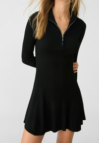 Robe noire côtelée avec col simulé à fermeture éclair. Manches longues, silhouette évasée, texture lisse et ourlet à hauteur du genou.