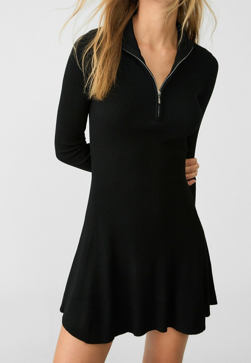 Robe noire côtelée avec col simulé à fermeture éclair. Manches longues, silhouette évasée, texture lisse et ourlet à hauteur du genou.