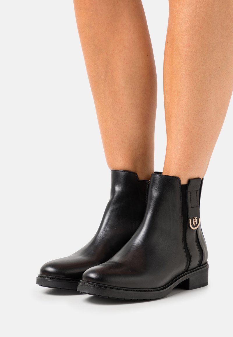 Bottines en cuir noir à talons bas, avec panneaux latéraux élastiques et boucle logo dorée, portées sur des jambes nues sur un fond uni.