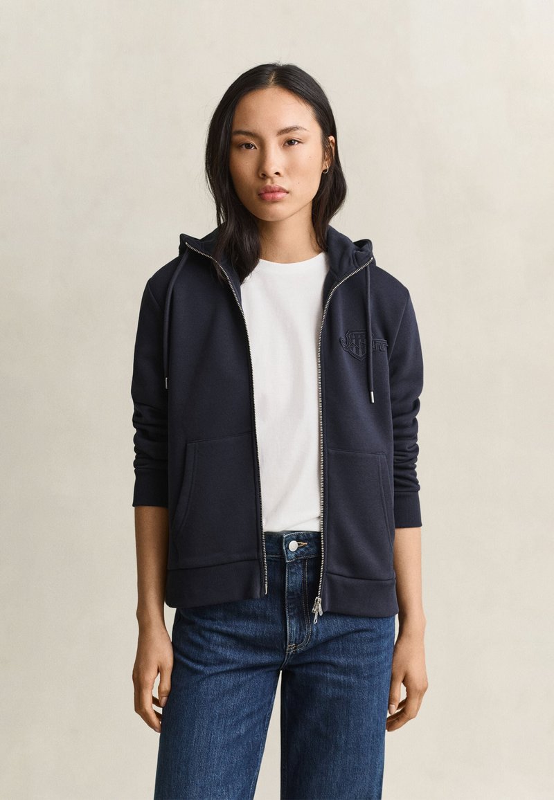 Jonge vrouw die een marineblauwe hoodie met rits draagt over een wit T-shirt en een blauwe spijkerbroek, staand tegen een effen achtergrond.