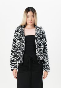 Zebra raštas dirbtinė vilna striukė su užtrauktuku, ilgomis rankovėmis ir gaubtu. Dėvima virš prigludusios juodos suknelės, turinčios tekstūruotą išvaizdą.