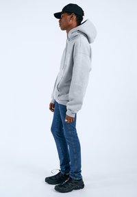 Felpa grigia con zip e un grande cappuccio, abbinata a jeans blu aderenti e sneakers nere. Il modello indossa un cappellino nero e un anello.
