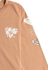 Dlhý rukáv tričko z jemnej broskyňovej látky s bielymi kvetinovými vzormi na hrudi a rukávoch, s hladkou textúrou a voľným strihom.