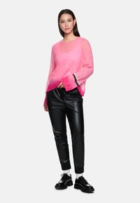 Rosa, halbtransparente Strickjacke mit weitem Ausschnitt und langen Ärmeln; kombiniert mit glänzenden schwarzen Jogginghosen und klobigen schwarzen Schuhen.