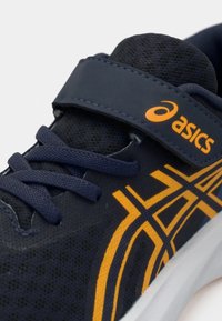 Μπλε αθλητικό παπούτσι με πορτοκαλί λεπτομέρειες, διαθέτει πλεκτό άνω μέρος, ιμάντα με Velcro και υφή με σχέδια. Τα κορδόνια είναι ορατά στο άνω μέρος.