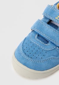 Pantof din piele întoarsă albastră, cu două curele velcro, vârf perforat și căptușeală albă din plasă. Prezintă cusături galbene contrastante și o talpă texturată.