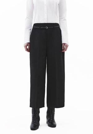 Personne portant un pantalon noir large et court avec une fine ceinture noire, une chemise blanche boutonnée et des bottines noires à lacets, positionnée de face.