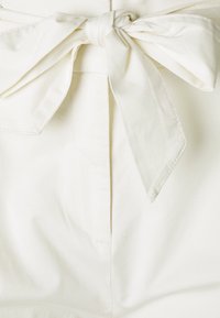 Tissu en coton blanc avec un nœud noué à la taille, présentant un devant plat et des détails cousus. Le matériau semble doux et lisse.