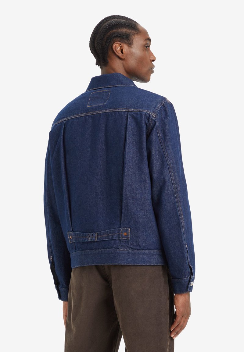 Mann mit geflochtenem Haar, der eine dunkelblaue Jeansjacke und braune Hose trägt, von hinten vor einem weißen Hintergrund gezeigt.