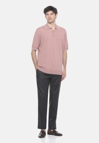 Uomo in piedi che indossa un polo a maniche corte rosa chiaro, pantaloni grigio scuro e mocassini marrone scuro su uno sfondo bianco semplice.