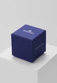 Swarovski blaue strukturiert Geschenkbox mit einem aufgedruckten Logo oben und der Webadresse an der Seite, präsentiert auf einem weißen Podest.