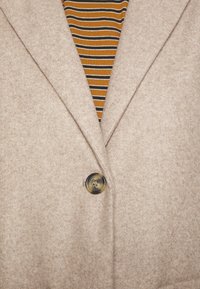 Béžový vlněný kabát s jedním knoflíkem zapnutým přes pletený top s hnědými, černými a bílými pruhy.