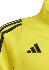 Geel sportjack van gestructureerde stof, met een zwart drie-strepenontwerp op de mouwen en een geborduurd zwart logo.