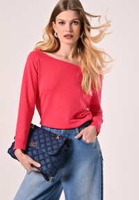 Roter Off-Shoulder-Pullover, blaue High-Waist-Jeans, dazu eine dunkelblaue gesteppte Handtasche mit goldener Verschluss- und Kettenakzenten.