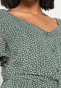 Vestido verde con escote en V, lunares blancos y detalles de botones. Tejido suave con mangas cortas y diseño ajustado en la cintura.