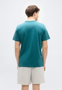 Μπλουζάκι από βαμβάκι σε τιρκουάζ χρώμα με στρογγυλή λαιμόκοψη και κοντά μανίκια, συνδυασμένο με γκρι αθλητικές βερμούδες. Το ύφασμα φαίνεται απαλό με άνετη, casual εφαρμογή.