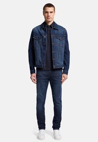 Giubbotto di jeans con due tasche sul petto, colore blu scuro e bottoni. Abbinato a jeans scuri e sneakers bianche. Design semplice e strutturato.