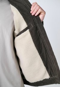 Chaqueta de pana marrón con forro de felpa crema, que presenta un bolsillo lateral y una textura acanalada, exhibida abierta con una mano agarrando el cuello.