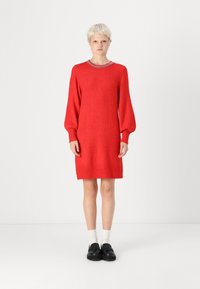 Robe en tricot rouge avec de longues manches bouffantes, un col rond, des poignets et un ourlet à côtes. Portée avec des chaussures noires et des chaussettes blanches.