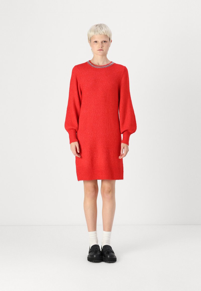 Robe en tricot rouge avec de longues manches bouffantes, un col rond, des poignets et un ourlet à côtes. Portée avec des chaussures noires et des chaussettes blanches.