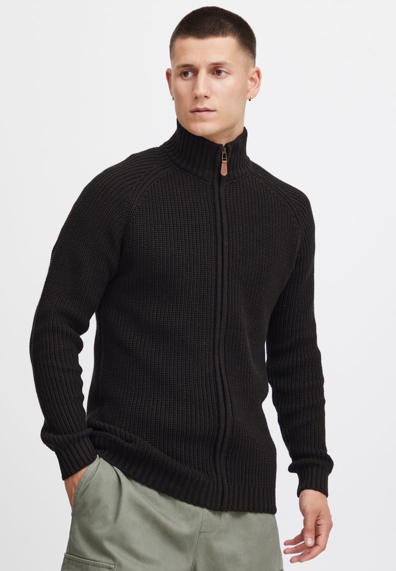 Schwarzer Strickpullover mit hohem Kragen und Reißverschluss, ausgestattet mit Rippstrick-Textur und langen Ärmeln. Zu olivgrünen Hosen getragen.