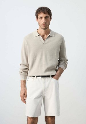 Hombre con camisa beige de manga larga y pantalones cortos blancos de pie contra un fondo sencillo y claro con una mano en el bolsillo.