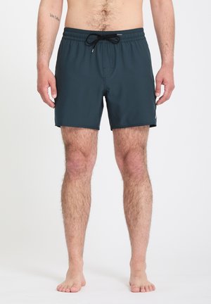 Homme portant un short de bain bleu foncé avec un cordon noir, debout pieds nus sur un fond blanc.