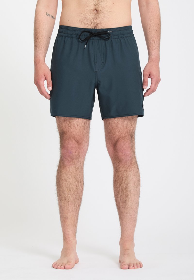 Homme portant un short de bain bleu foncé avec un cordon noir, debout pieds nus sur un fond blanc.