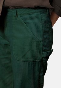 Groene ribfluweel broek met verticale ribbels, voorzien van een zijzak en een lusdetail op het been. Het materiaal heeft een zachte textuur.
