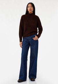 Brun tekstureret striktrøje med høj hals, parret med vidde ben mørkeblå denimjeans og sorte chunky sko. En simpel og afslappet outfit.