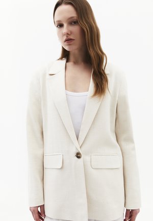 Jonge vrouw met lang bruin haar die een oversized crèmekleurige blazer draagt over een wit topje, staand tegen een effen witte achtergrond.