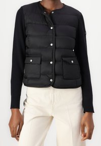 Zwarte puffer gilet met voorste drukknoopsluitingen, twee opgestikte zakken en gebreide lange mouwen. Glad textuur met een getailleerde silhoeet.