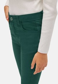 Los pantalones de algodón verdes presentan un diseño ajustado con bolsillos frontales y una textura suave, acompañados de una camiseta de manga larga de color claro y acanalada.