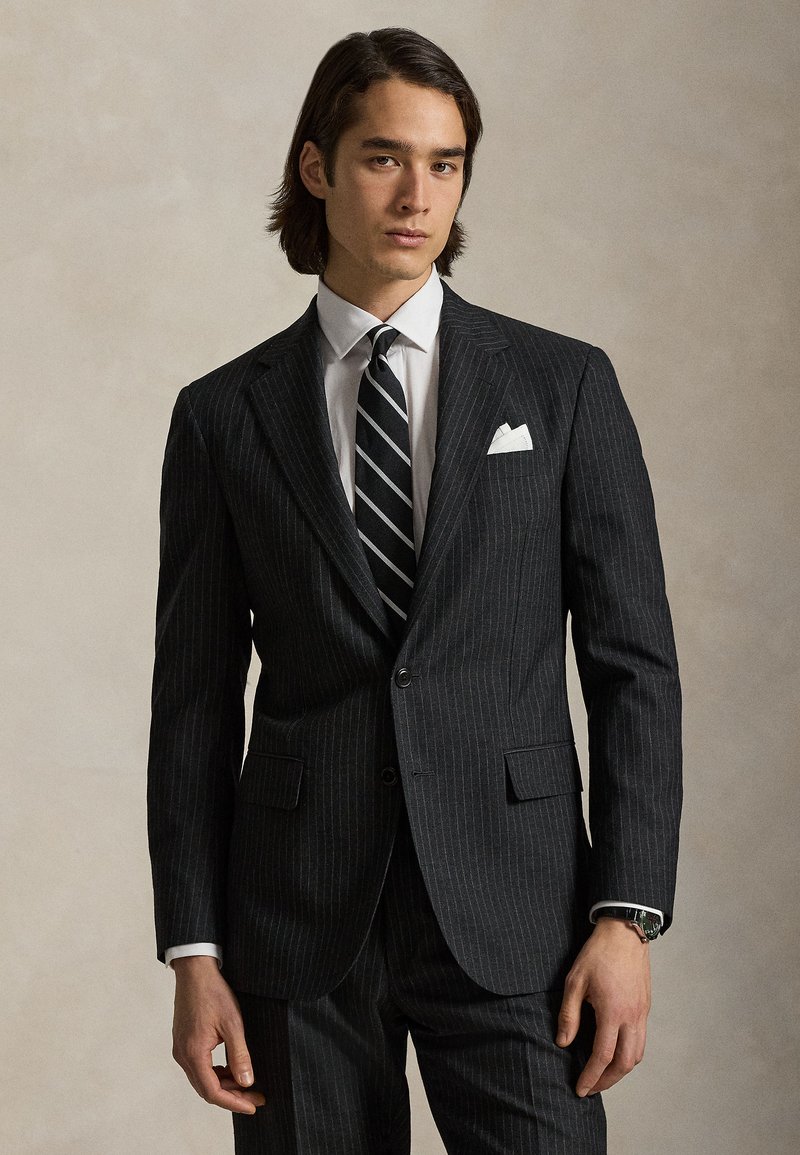 Νεαρός άνδρας φοράει σκούρο κοστούμι με ρίγες, λευκό πουκάμισο, γραμμωτή γραβάτα, λευκή μαντήλι τσέπης και ρολόι χειρός, στέκεται μπροστά σε απλό φόντο.