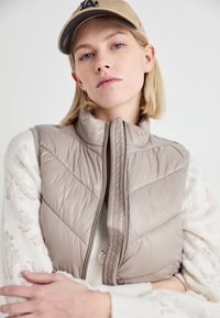 Gilet rembourré beige clair avec un col haut, un matelassage vertical et une fermeture éclair à l'avant. Porté par-dessus un pull blanc texturé, assorti avec une casquette beige.