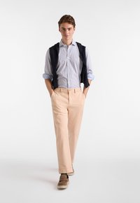 Pantalones rosa pálido, camiseta a rayas azules y chaleco azul oscuro, caminando en un fondo neutral. Calzado informal completa el conjunto.