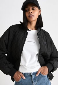 Giacca bomber nera imbottita con polsini a costine, chiusura con zip e dettaglio del marchio, indossata sopra una t-shirt bianca e jeans blu. Cappellino nero incluso.