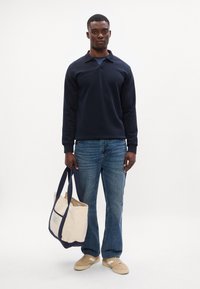 Tengeri kék hosszú ujjú póló, kék farmernadrág, bézs sportcipő és egy krém színű táska sötétkék díszítéssel és kiszabott anyaggal. Egyszerű háttér.