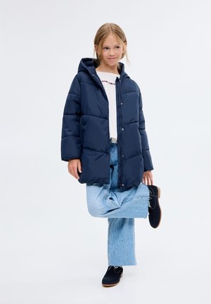 Giacca puffer blu navy con cappuccio, caratterizzata da pannelli cuciti, bottoni a pressione e una vestibilità comoda. Indossata sopra a una maglia bianca e jeans azzurri.