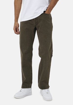 Homme portant un pantalon vert foncé coupe décontractée, des baskets blanches et une chemise blanche, une main dans la poche, sur un fond uni.