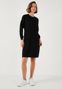 Black long-sleeve dress with a relaxed fit, made of soft fabric. Features a round neckline and falls above the knee, paired with white sneakers.