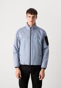 Light blue jacket with a stand-up collar, front zip, and black accents. Features elastic cuffs and a zippered pocket on the sleeve.